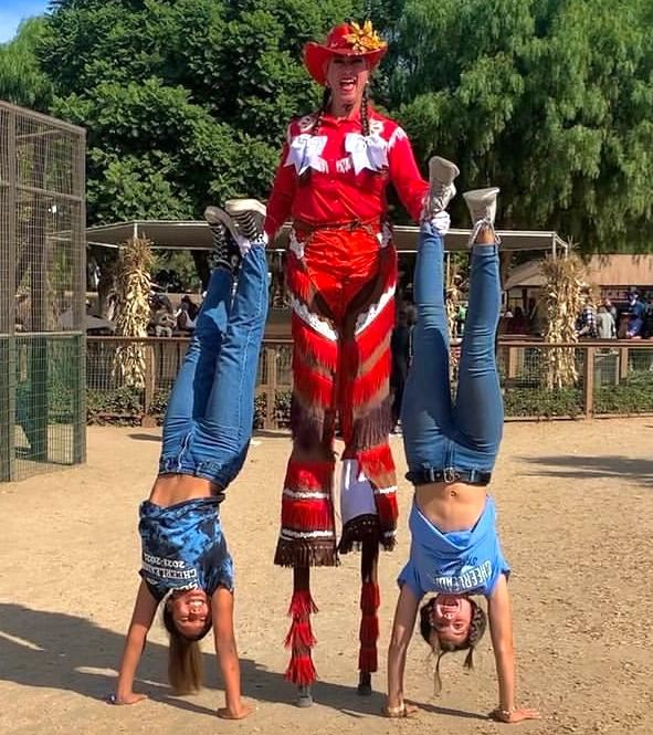 Woman in red on stilts holding two people upside down doing handstands by their foot