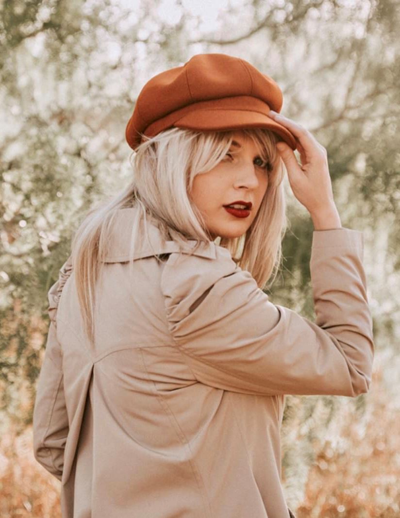 A blonde woman looking backward to the camera with a brown hat, beige jacket and Taylor Swift iconic red lipstick