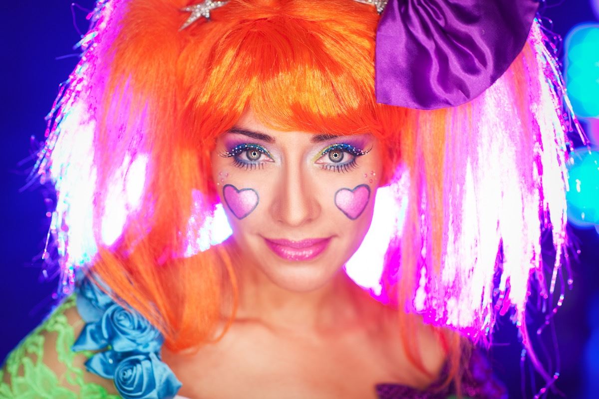 Twinkle Time looking into the camera. She has big orange hair with a purple bow and sparkly makeup