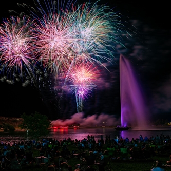 Fireworks are exploding over the lake at Fountain Park. The fountain is lighted in purple in the photo.