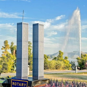 Small American flags line the ground in front of a small replica of the World Trade Towers with the town's fountain in the background.