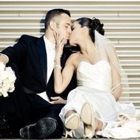 A man in a black suit and a woman in a wedding dress share a kiss while seated on the floor with their backs to the wall.