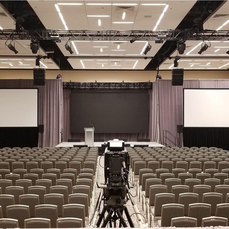 Large open room with a stage in between two large projection screens. Multiple rows of chairs are in front of the stage.