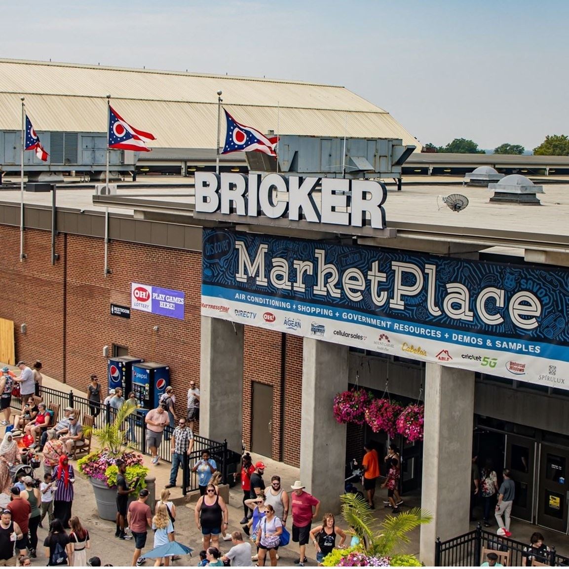 Building with sign that reads Bricker MarketPlace with flags on the top of the building and people waking in front of building.