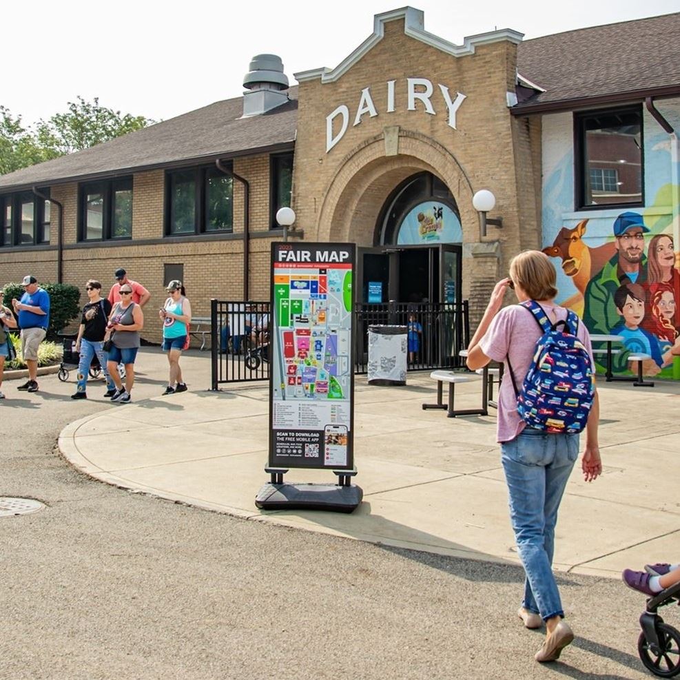 Person walking with backpack toward printed vertical map in front of the Dairy Products Building at the Ohio State Fair.