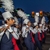 Flute players and a clarinet player in a marching band, with lights on the instruments, wearing red, white & blue uniforms with tall white feather plumes in their hats