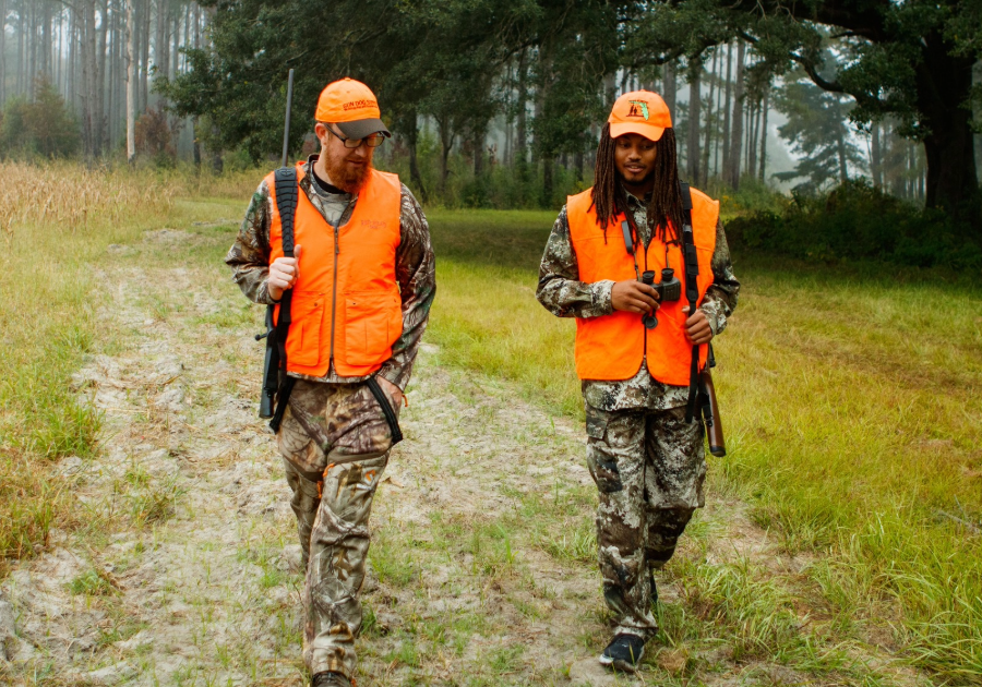 2 men in camouflage and an orange vest  walking in the forest